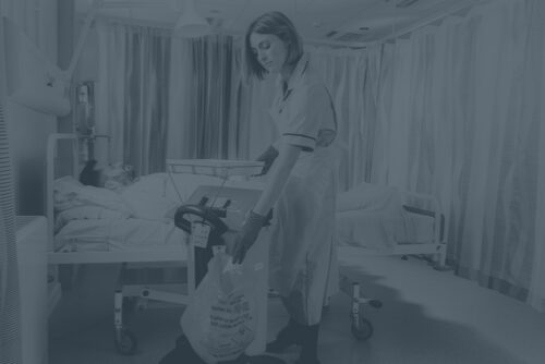 Female nurse disposing of medical waste in Sharpsmart container next to patient in a hopsital room